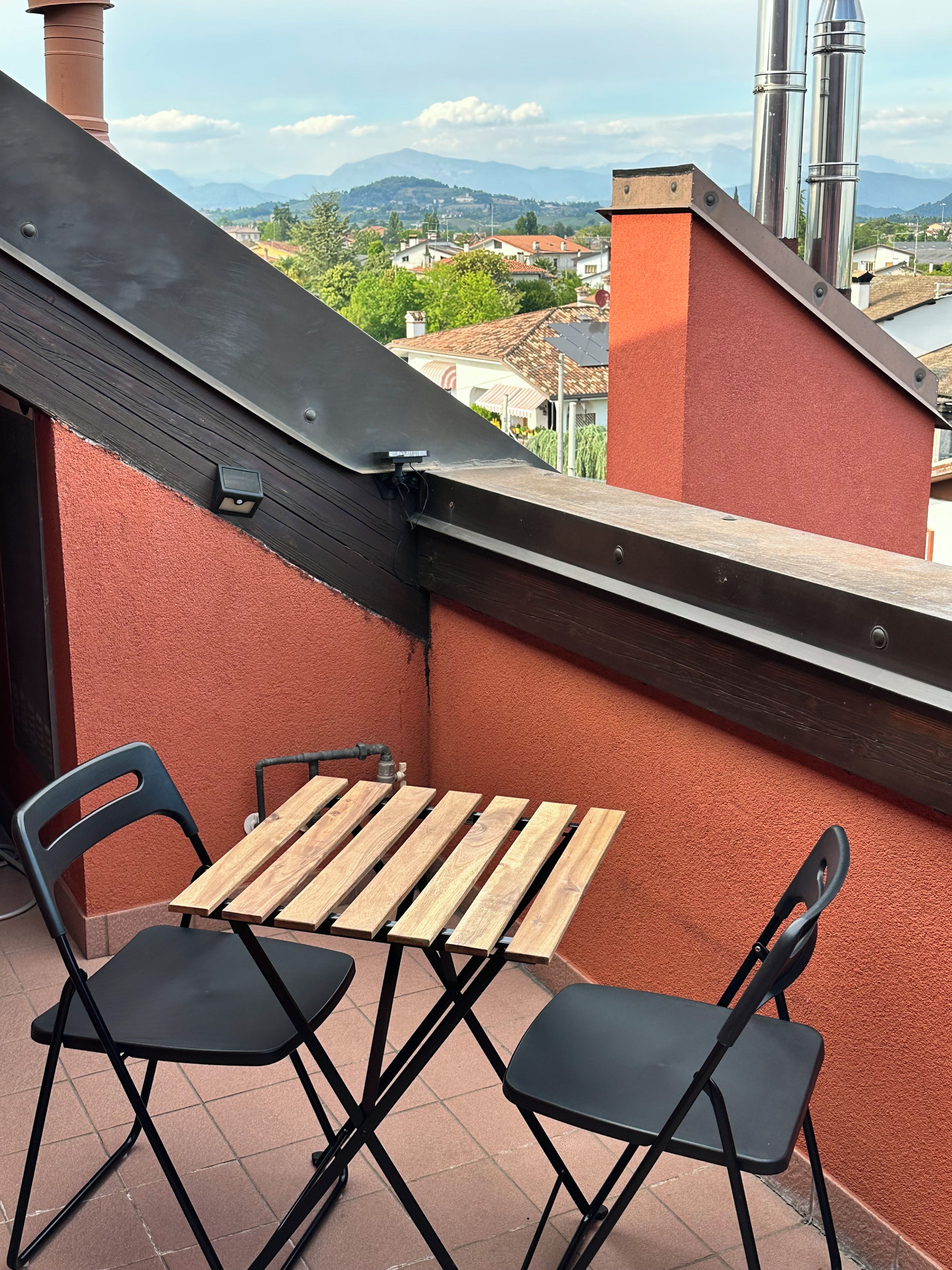 Un petit espace extérieur aménagé avec une table en bois et deux chaises, offrant une vue sur un quartier résidentiel avec des maisons et des montagnes à l’horizon.