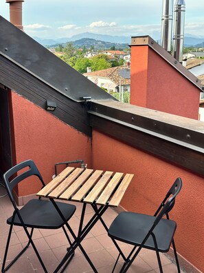 Een klein buitenzitje met een houten tafel en twee stoelen, met uitzicht op een woonwijk met huizen en bergen in de verte.