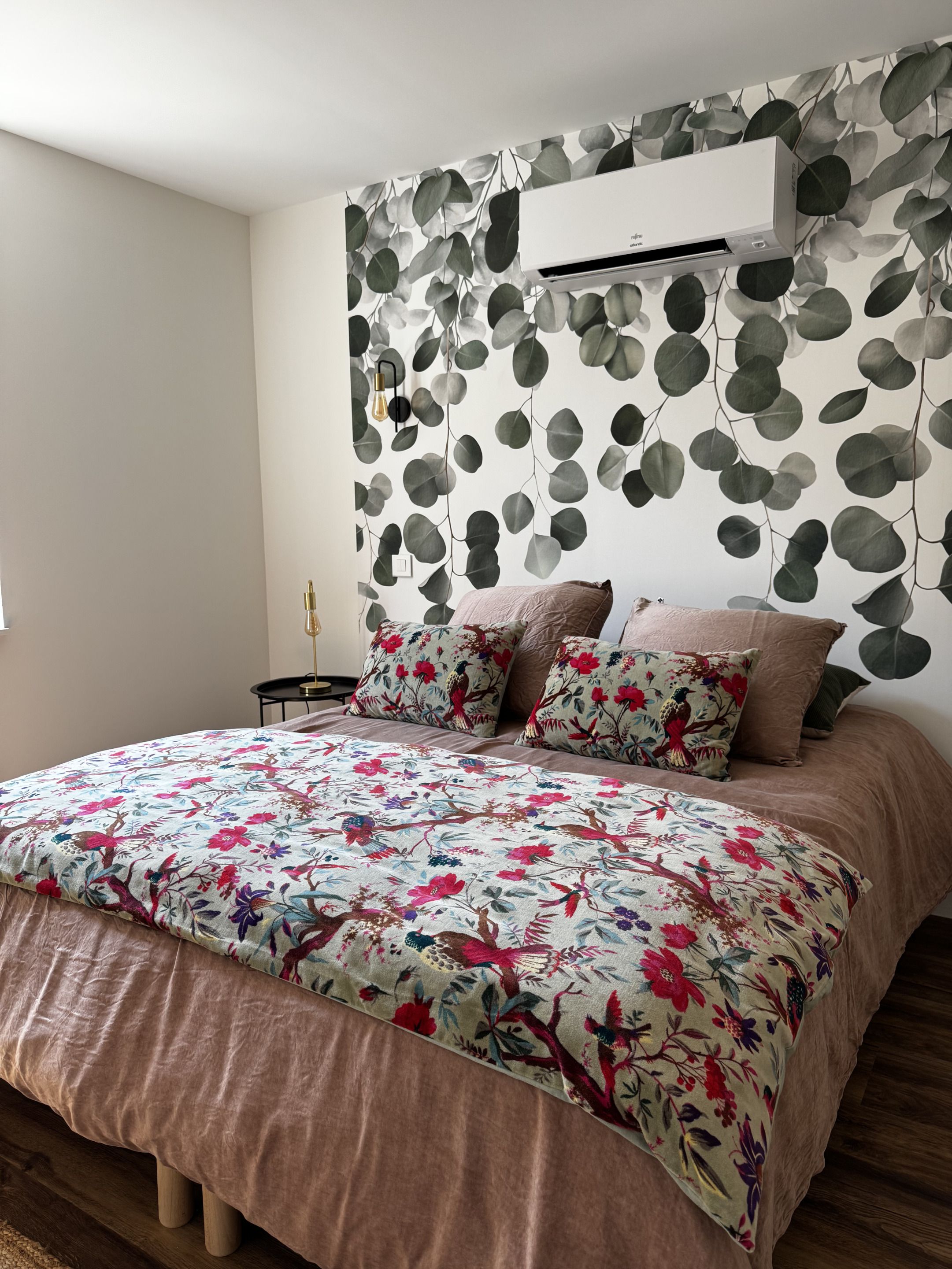 A bedroom with a bed, floral bedding, a wall-mounted air conditioner, and a wall with a leafy pattern.