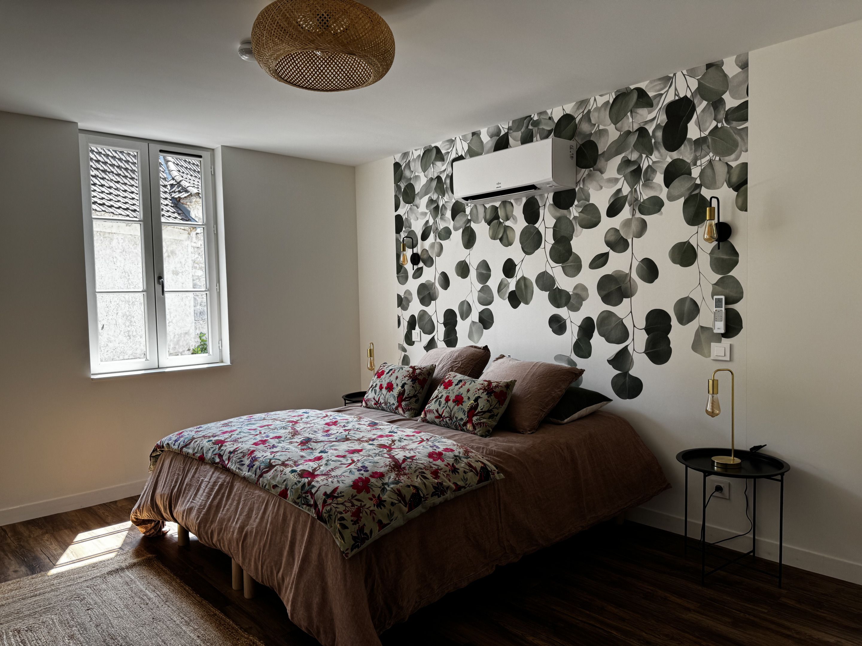 A bedroom with a bed, a window, a wall-mounted air conditioner, and a small bedside table.