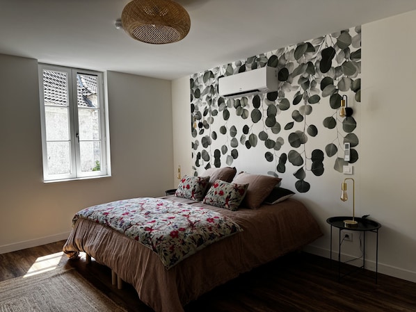 A bedroom with a bed, a window, a wall-mounted air conditioner, and a small bedside table.