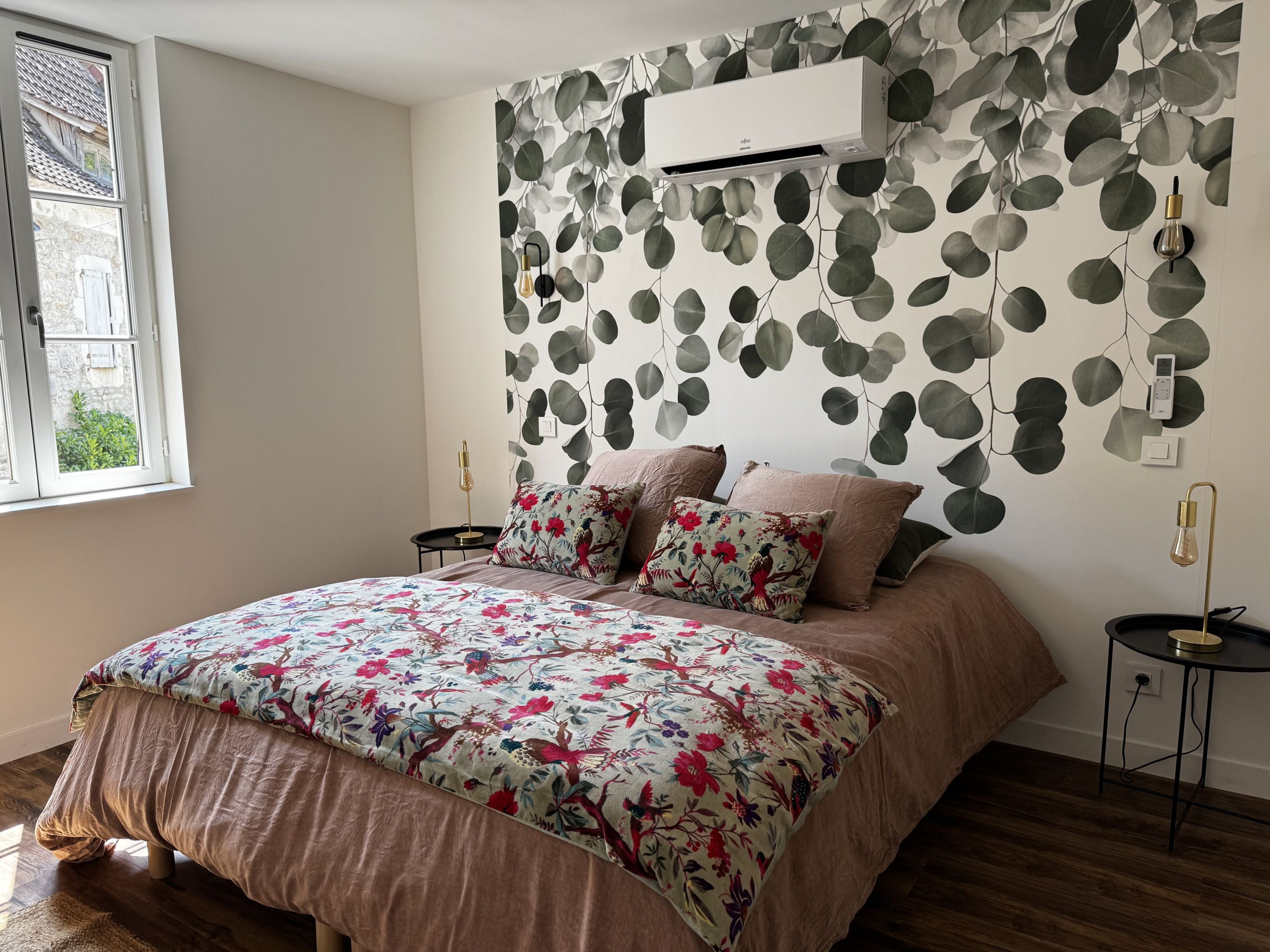 A bedroom with a bed, a window, an air conditioner, and a wall with a leaf pattern.