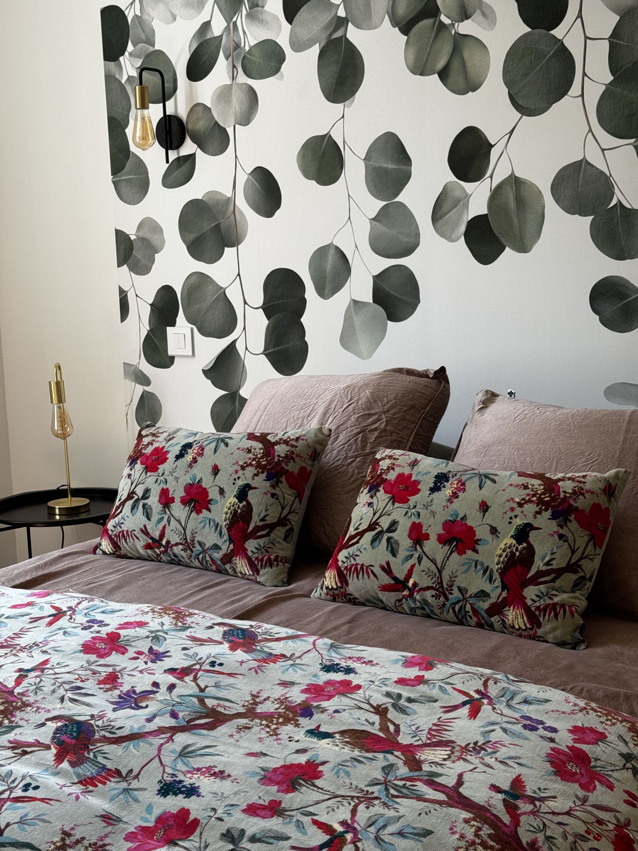 A bedroom with a bed, two pillows with floral patterns, a wall-mounted light fixture, and a wall with a leaf pattern.