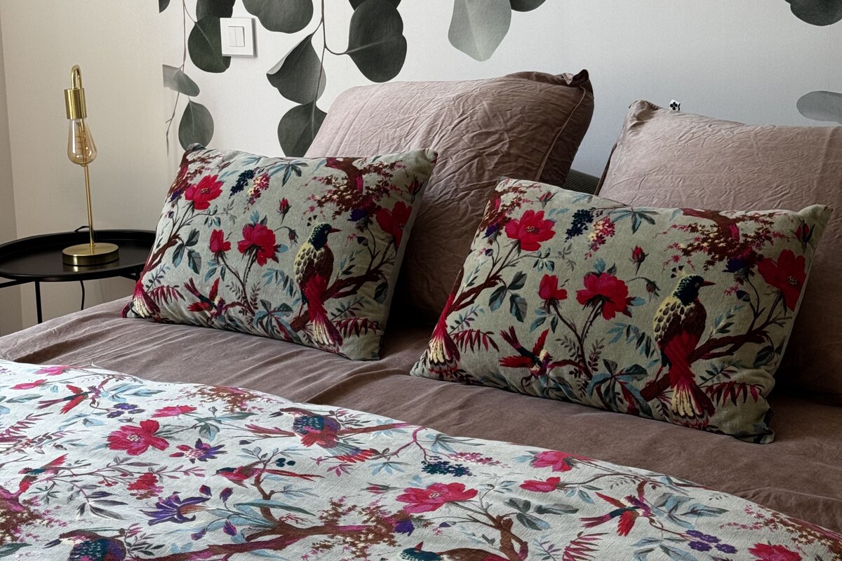 A bedroom with a bed, two pillows with floral patterns, a wall-mounted light fixture, and a wall with a leaf pattern.