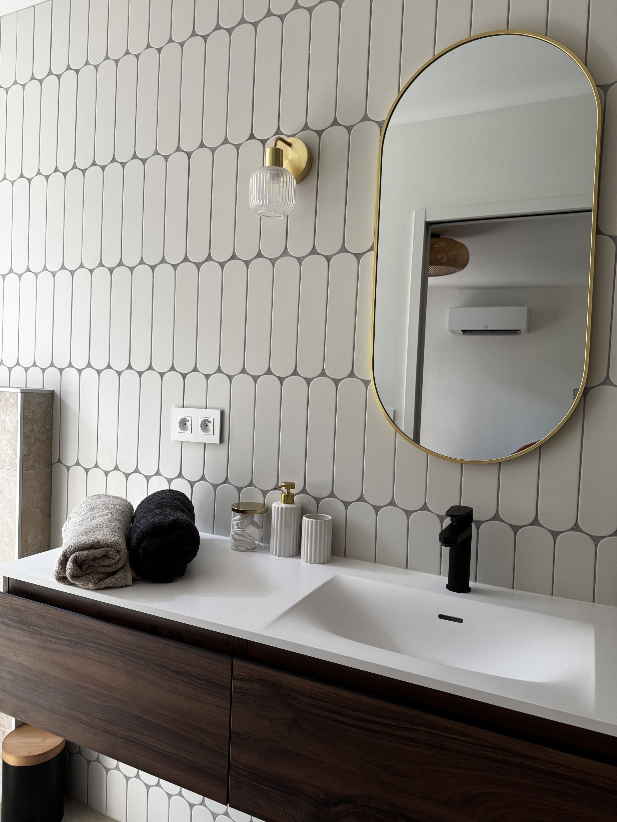 Un baño con pared de azulejos blancos, un espejo redondo con marco dorado, un lavabo blanco, un mueble de tocador de madera oscura y dos toallas.