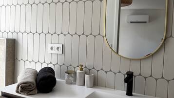 Un baño con pared de azulejos blancos, un espejo redondo con marco dorado, un lavabo blanco, un mueble de tocador de madera oscura y dos toallas.