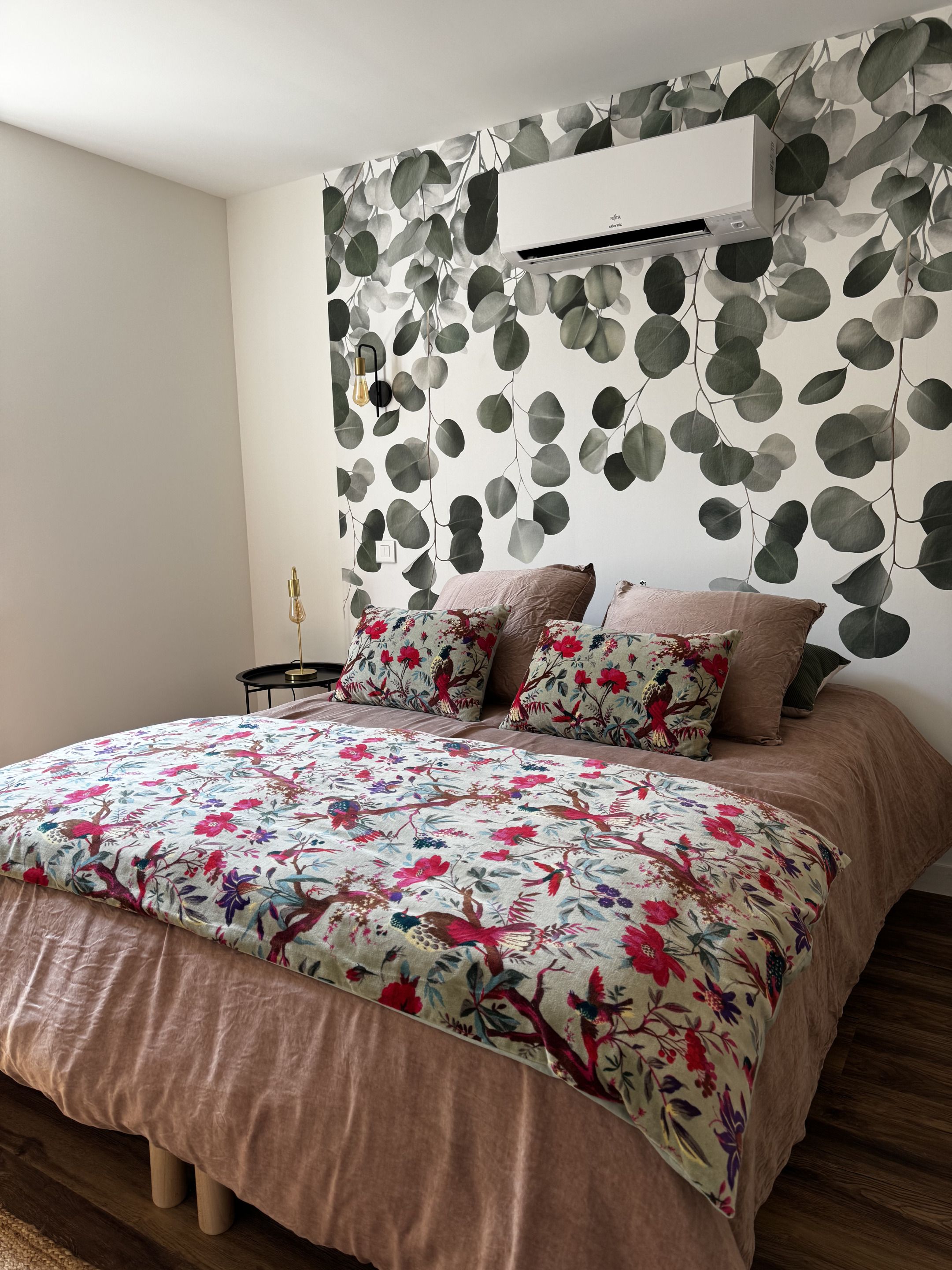 A bedroom with a bed, pillows, a wall-mounted air conditioner, and a wall with a leaf pattern.