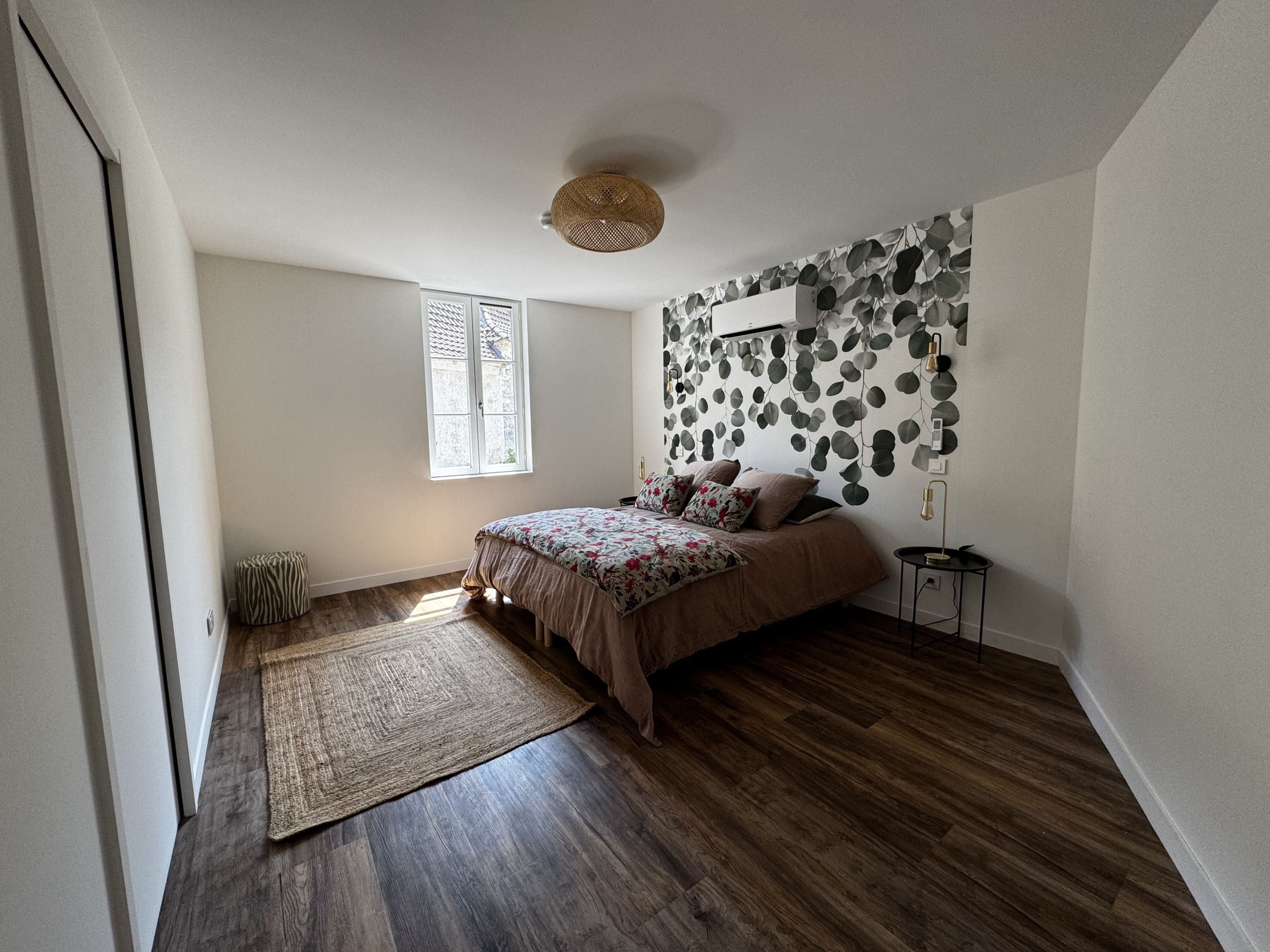 A bedroom with a bed, a window, a wall-mounted air conditioner, a small bedside table, and a woven floor lamp.