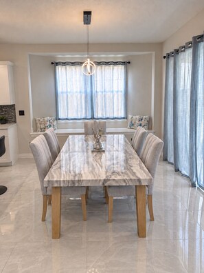 A dining area with a marble table, cushioned chairs, a pendant light, and a window with curtains. - Luxury Spacious 6-Bedroom Pool Home Near Disney Sleeps 18 (Haines City)