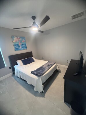 Ein Schlafzimmer mit einem Bett, Deckenventilator, einem Gemälde, einem Schreibtisch und einem Fernseher.