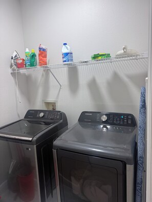 A laundry area with two front-loading washing machines and a shelf above containing various cleaning products. - Luxury Spacious 6-Bedroom Pool Home Near Disney Sleeps 18 (Haines City)