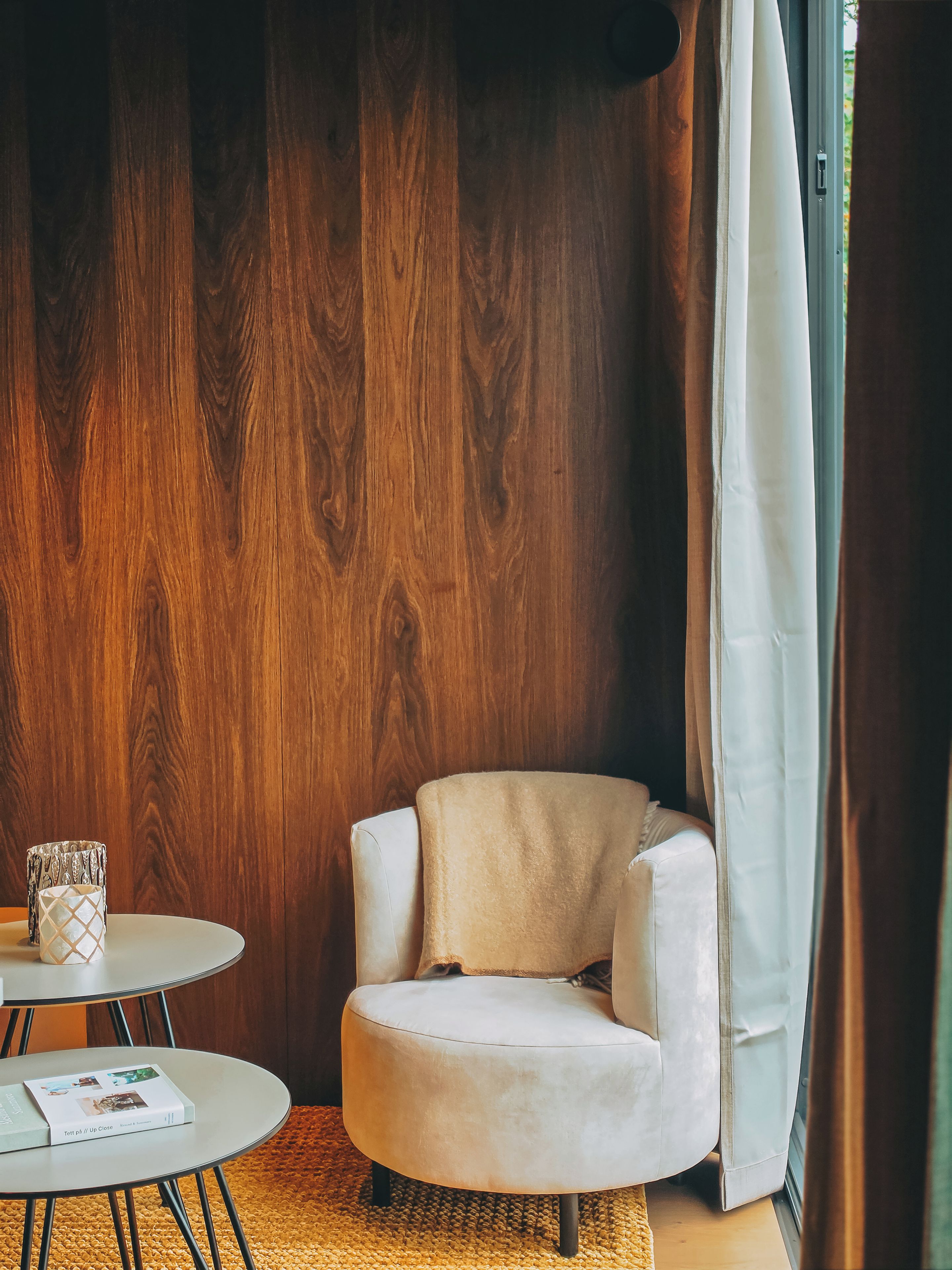 Een kamer met een houten wand, een ronde tafel met een boek, een fauteuil en een raam met gordijnen.