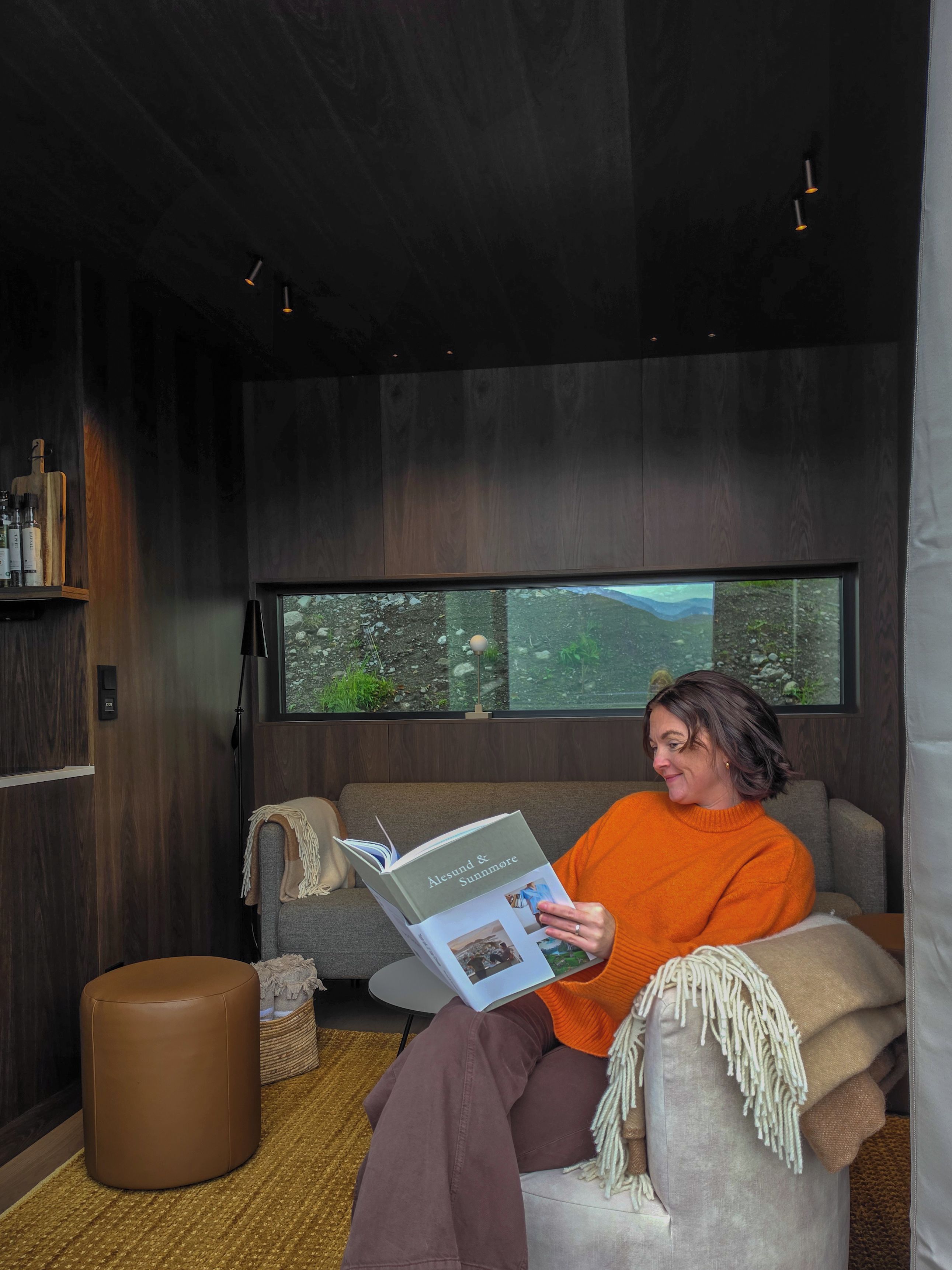 Une personne est assise sur un canapé, en train de lire un livre, dans une pièce dotée d’une grande fenêtre, d’un mur en bois et d’un petit pouf rond.