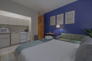 A bedroom with a bed, a green sofa, a desk, and a kitchen area with cabinets and appliances. - Polished Studio w/ Brick Accents (Boston)