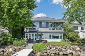 A two-story house with a covered patio, a stone retaining wall, and a view of the water. - Luxury Lakeside Oasis- 4 Bed / 3 Bath Lake Home (Middleton)