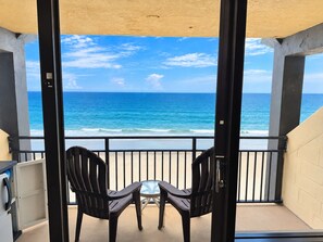 Un balcon avec deux chaises et une petite table, donnant sur une plage, l’océan et un ciel dégagé.