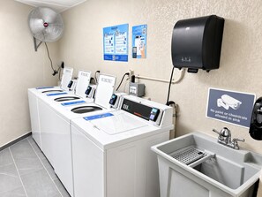 Une buanderie équipée de plusieurs machines à laver, d’un sèche-mains et d’un panneau indiquant qu’il est interdit de verser de la peinture ou des produits chimiques dans l’évier.