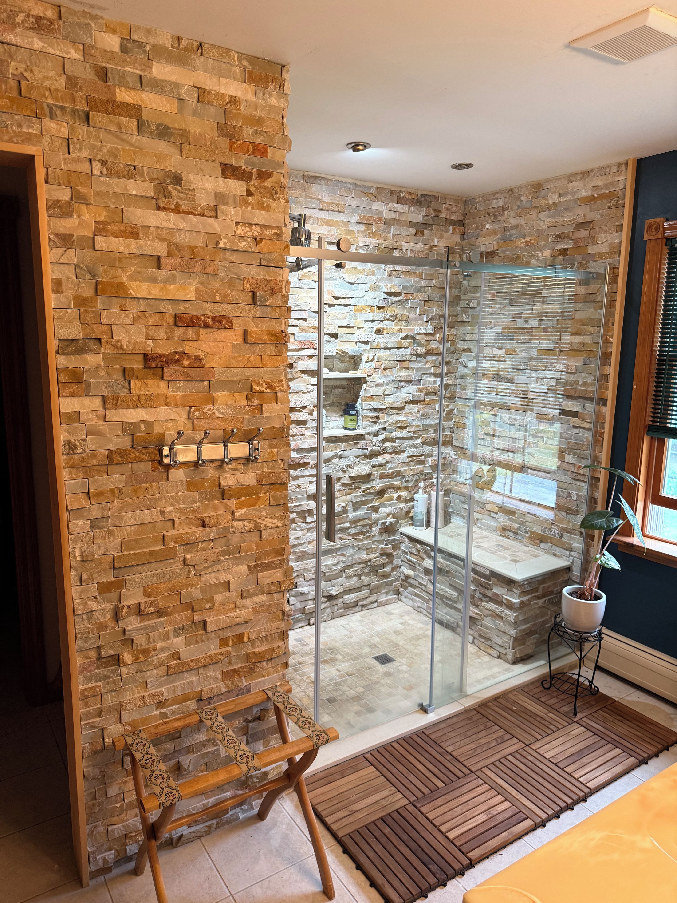 A bathroom with a stone wall, a glass shower enclosure, and a wooden bench.