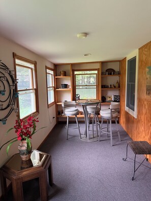 A compact kitchen with a dining area, wooden shelves, and a window with a view of trees. - Serene 3-bedroom 3-full BR house with Heat in charming Manchester Center, VT. (Manchester)