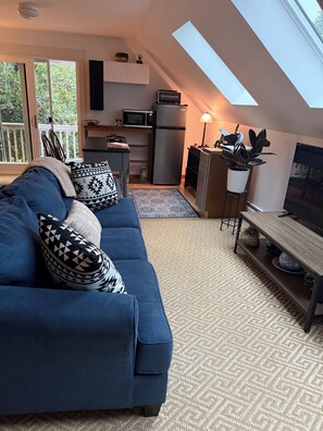 A modern living room with a blue sofa, patterned pillows, a TV, and a kitchen area with appliances. - Serene 3-bedroom 3-full BR house with Heat in charming Manchester Center, VT. (Manchester)