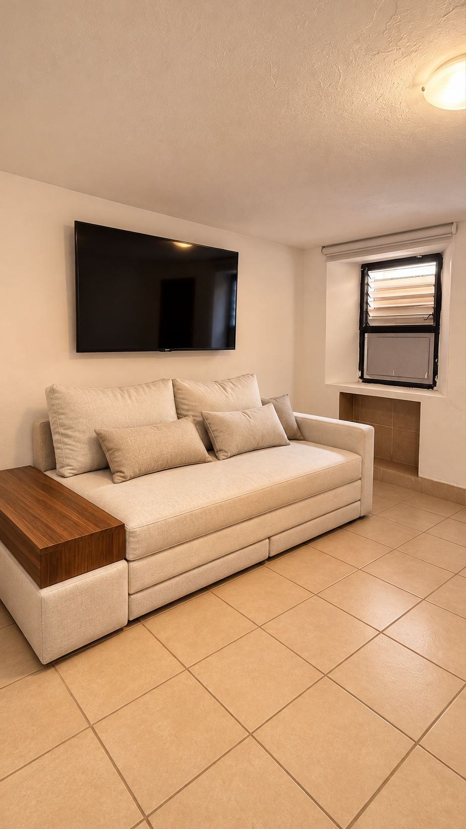 A living room with a sofa, a flat-screen TV, a window with blinds, and a tiled floor.