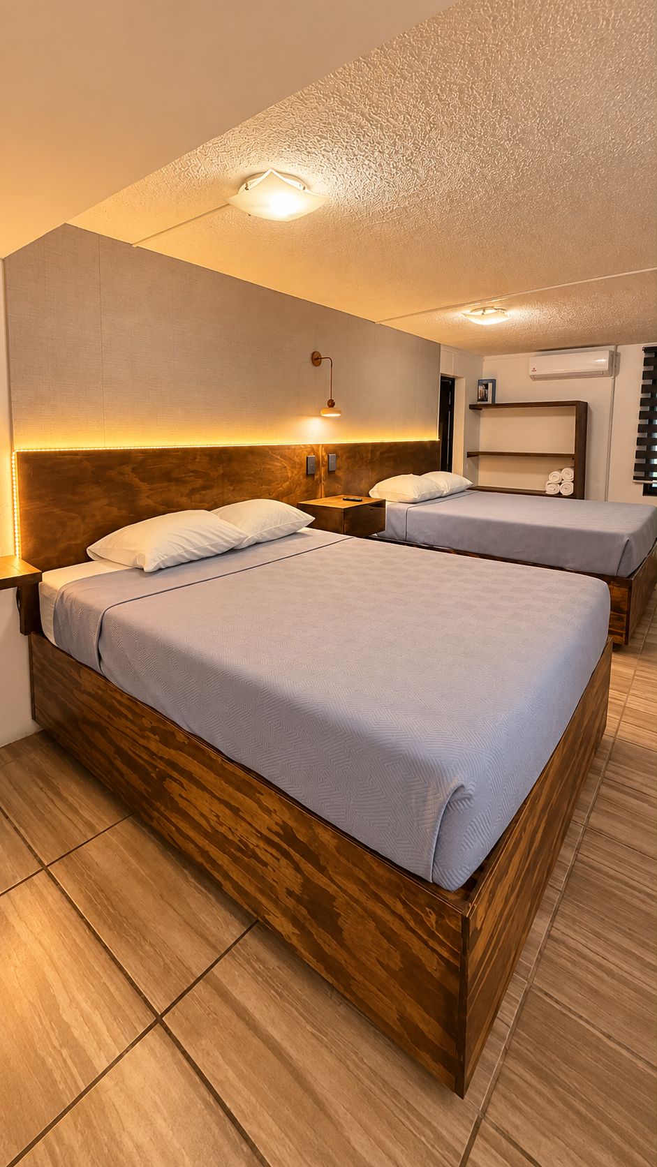 Habitación de hotel con dos camas, cabeceros de madera, una mesita de noche, un estante en la pared y una lámpara en el techo.