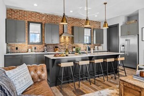A modern kitchen with a brick wall, grey cabinets, a central island with bar stools, and a wooden dining table. - Rooftop skyline views, walkable WeHo house, sleeps 10 near Geodis Park (Nashville)