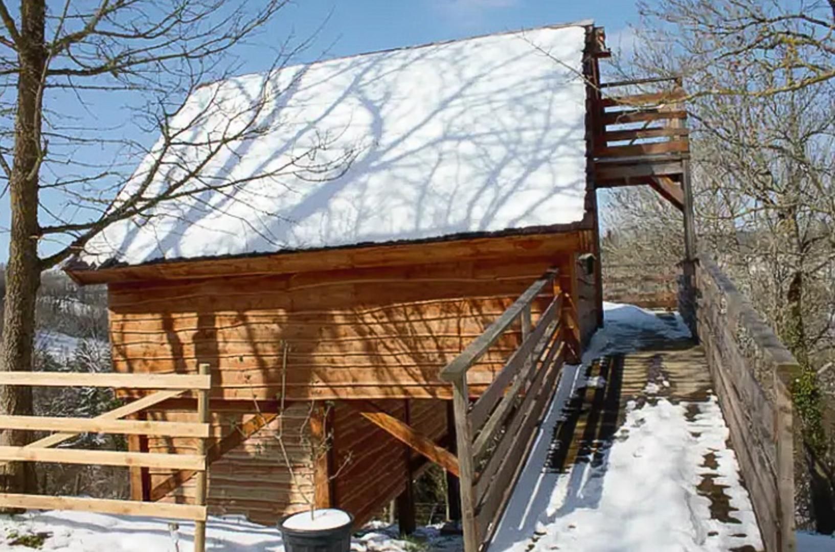 A wooden structure with a snow-covered roof, a wooden staircase leading up to a door, and a wooden fence surrounding the area.
