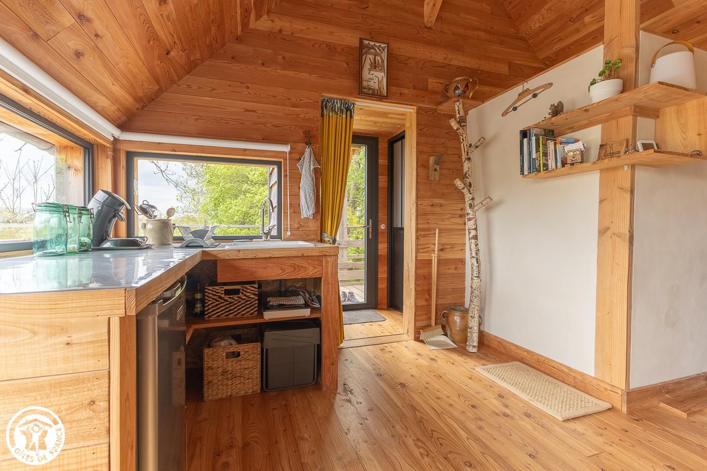 A wooden interior with a kitchen area, including a sink, shelves, and a window with a view.