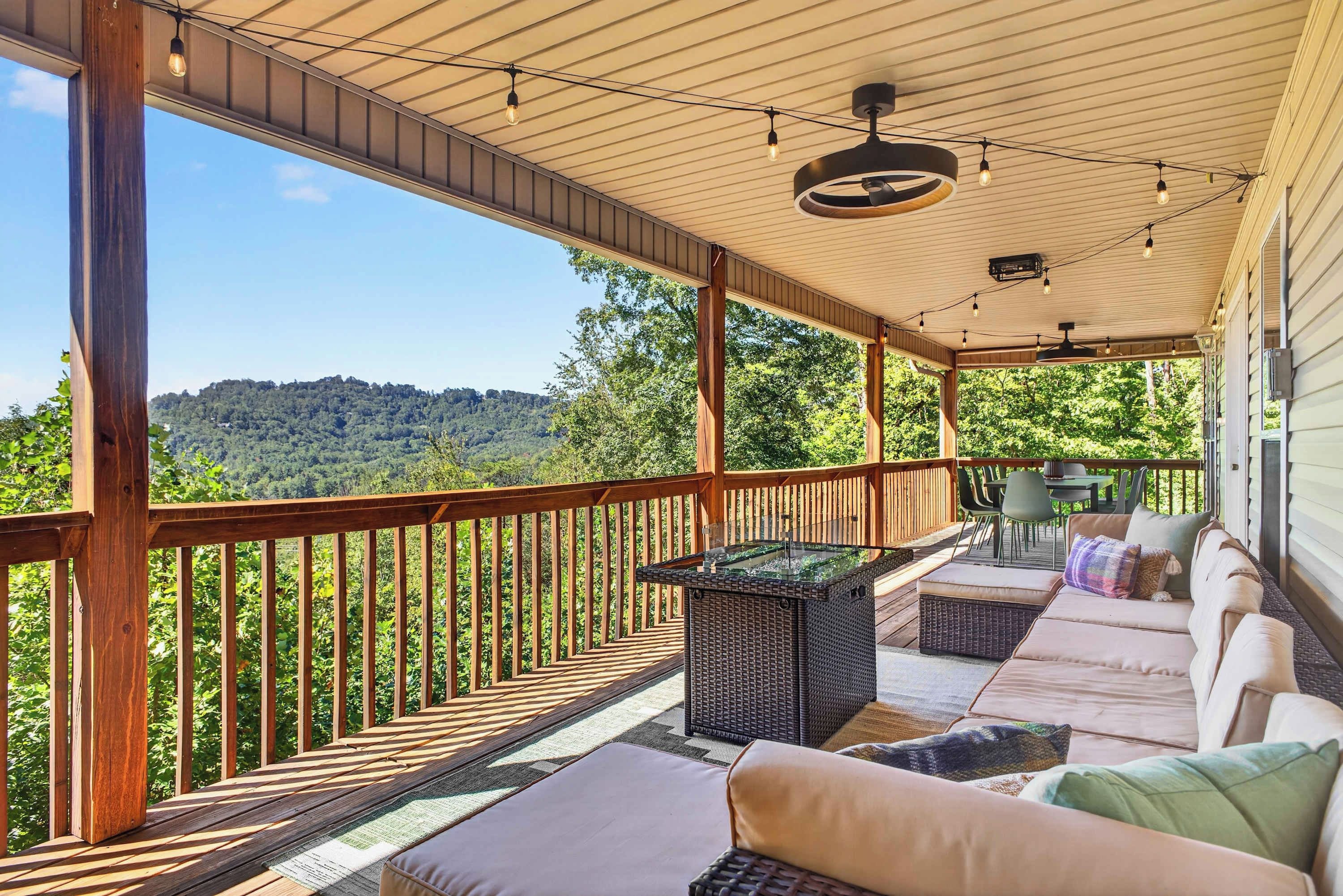 A covered outdoor deck with a view of a forested hill, wooden railings, and outdoor furniture.