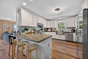 A modern kitchen with white cabinets, a central island with bar stools, stainless steel appliances, and a large window providing natural light. - Wineberry Ridge | Hot Tub • Sauna • Grill • Views (Asheville)