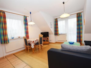 A living room with a sofa, a dining table with chairs, a TV, and a window with curtains. - Wunderschönes Appartement in Büsum mit Grill by Interhome (Büsum)
