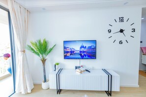 Ein modernes Wohnzimmer mit einem an der Wand befestigten Flachbildfernseher, einem weißen Tisch mit einem dekorativen Gegenstand, einer Topfpflanze und einer großen Wanduhr.