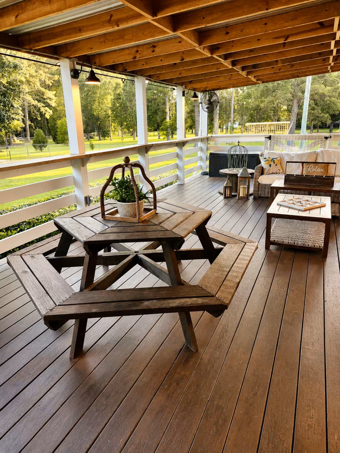 A covered outdoor area with wooden picnic tables and benches, a seating area with cushions, and a view of a green landscape.