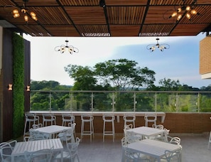 Un patio al aire libre con mesas y sillas, techo de madera y vista a árboles.