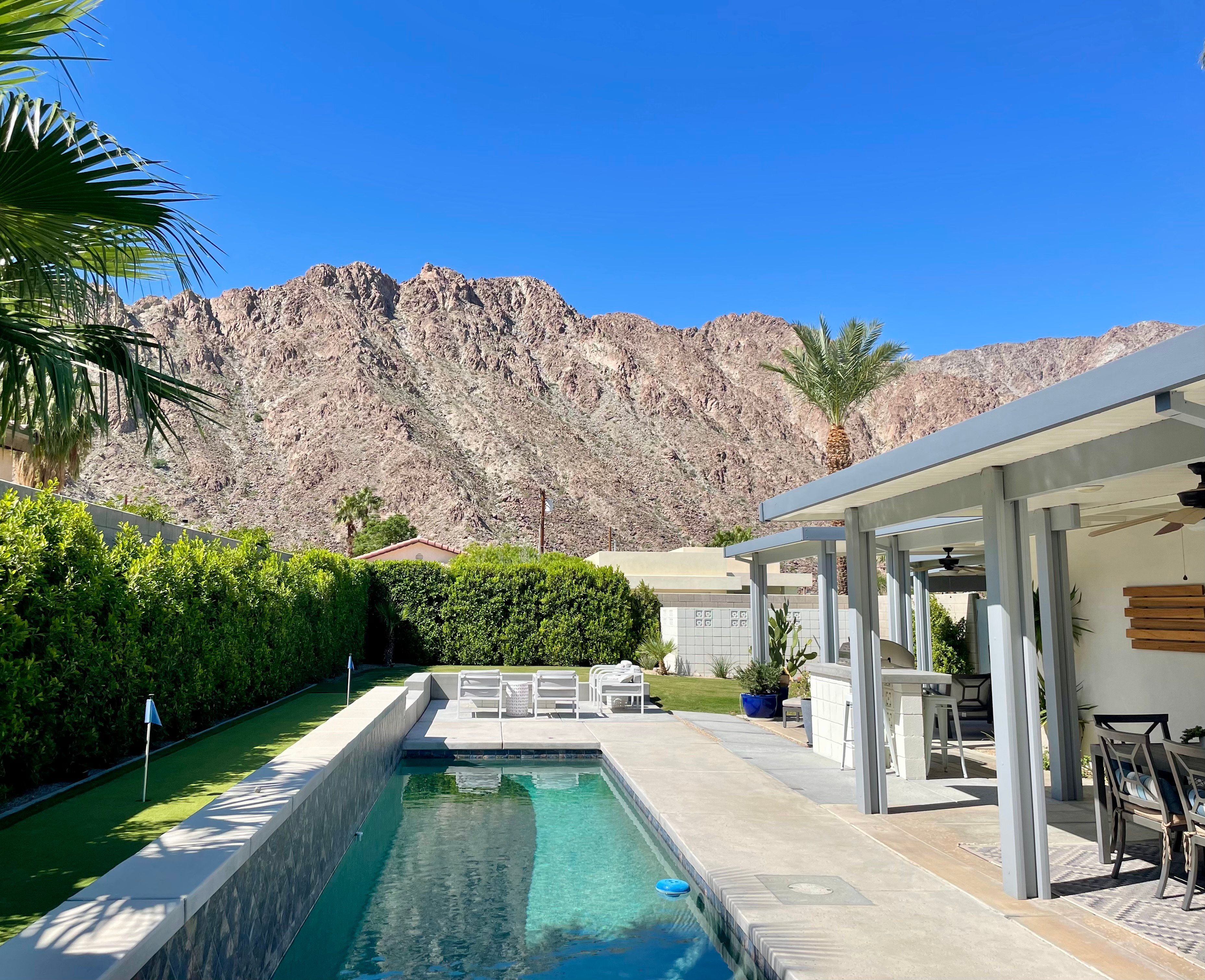 A swimming pool with a mountain backdrop, a covered patio area with seating, and a well-maintained hedge.