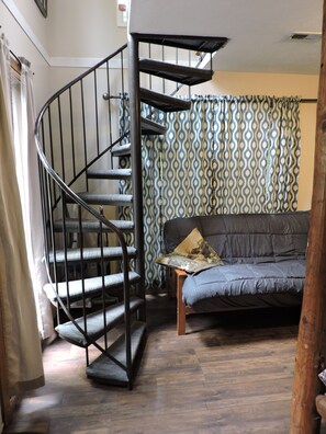 Eine Wendeltreppe mit Metallgeländern führt zu einer oberen Etage, ein gemustertes Vorhang, ein dunkelgraues Sofa mit Kissen und ein Holzboden.