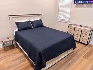 A neatly made bed with a dark quilt, a bedside table with a remote control, and a wooden nightstand. - New 3BR/2.5BA Near LBJ Hospital - Corporate & Extended Stays (Houston)