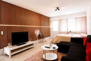 Una camera d'hotel moderna con televisione a schermo piatto, una ventola da terra, un divano, un tavolino rotondo e un letto con coperta beige.