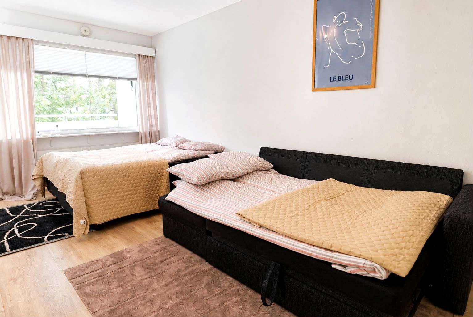 A room with two beds, a black headboard, a window with curtains, and a framed artwork on the wall.