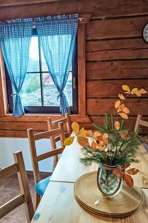 A wooden cabin interior with a dining table, chairs, a window with checkered curtains, and a vase with dried leaves.
