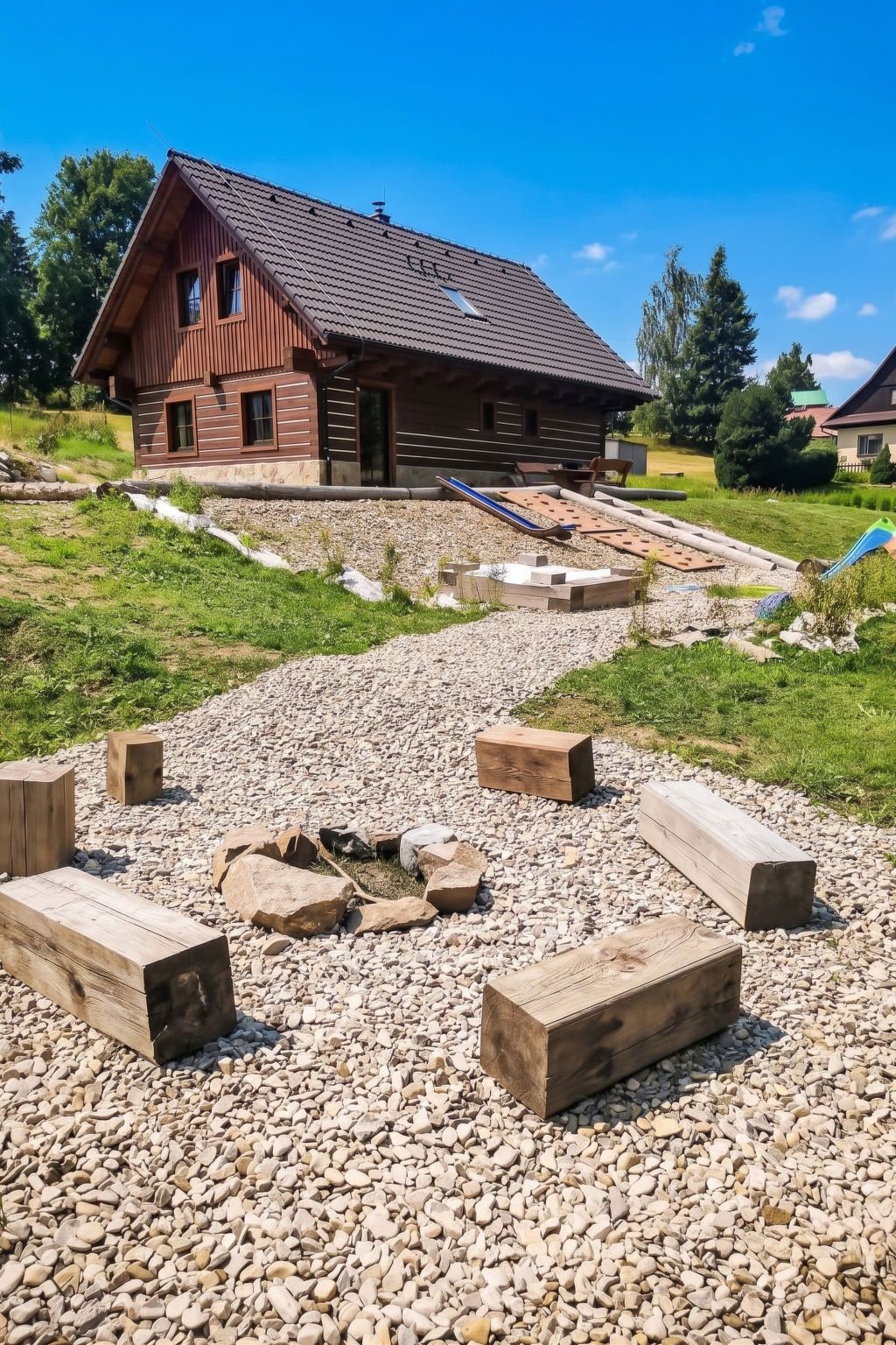 A wooden cabin with a sloped roof, surrounded by a gravel area with wooden benches and a small fire pit.