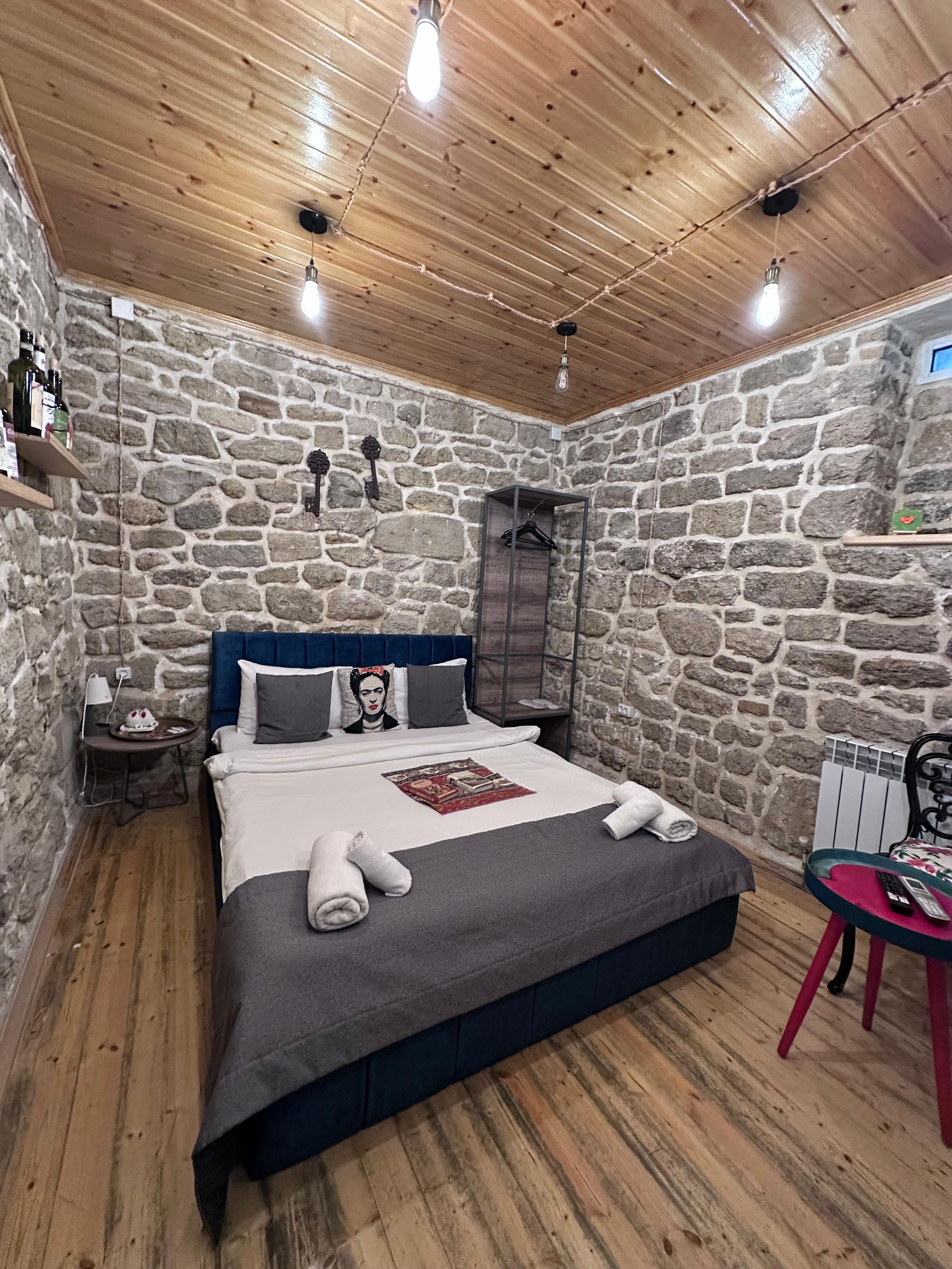 A bedroom with a stone wall, a bed with a blue headboard, a wooden floor, and a small table with a remote control.