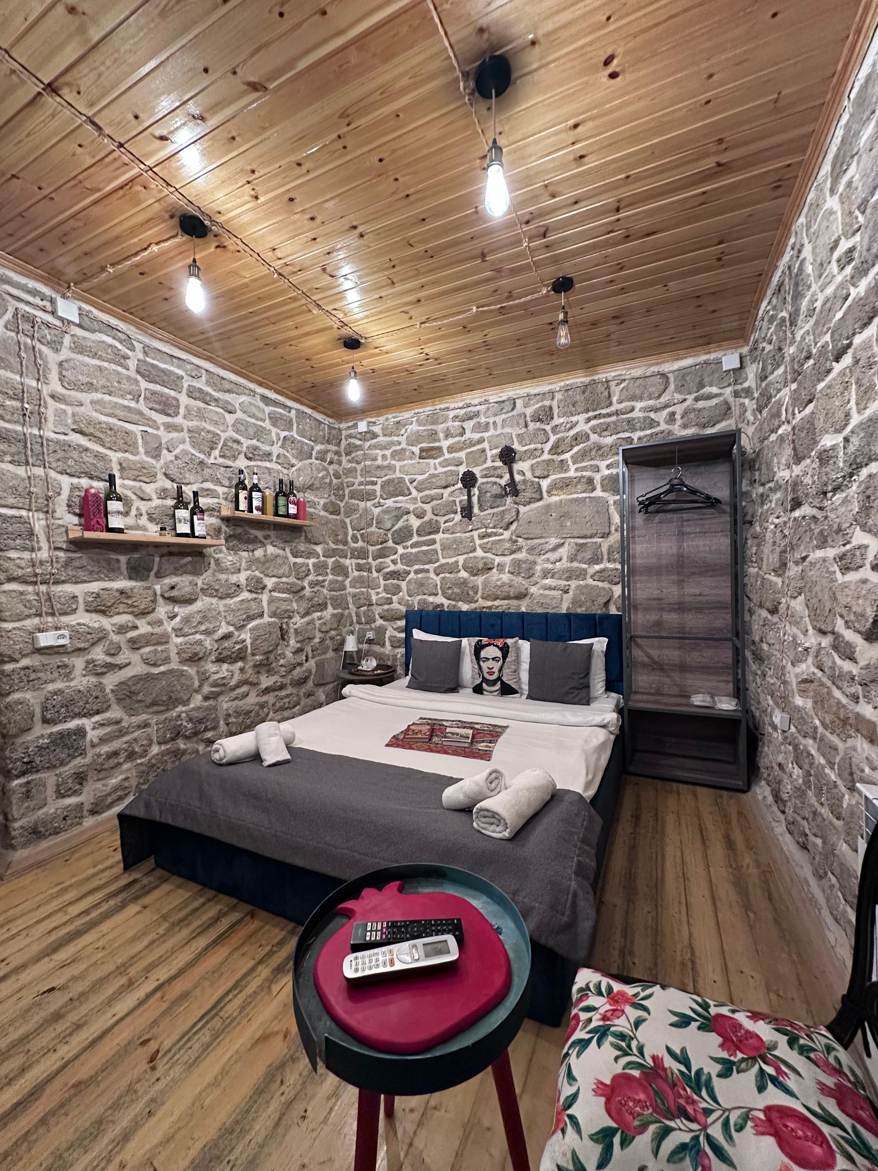 Ein Zimmer mit Steinwand, Holzbalkendecke, einem Bett mit Kissen, einem Nachttisch, einem hängenden Kleiderständer und einem kleinen Tisch mit Fernbedienung.