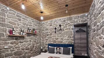 Ein Zimmer mit Steinwand, Holzbalkendecke, einem Bett mit Kissen, einem Nachttisch, einem hängenden Kleiderständer und einem kleinen Tisch mit Fernbedienung.