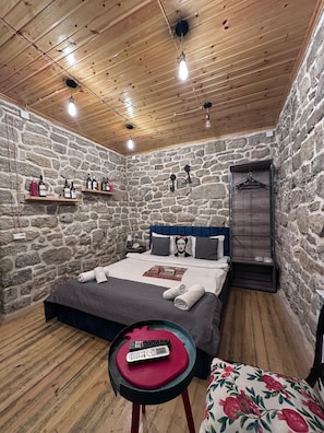Een kamer met een stenen muur, houten plafond, een bed met kussens, een nachtkastje, een kapstok en een klein tafeltje met een afstandsbediening.
