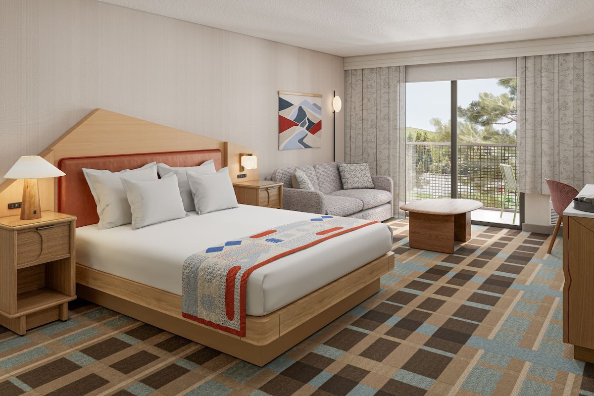 Een hotelkamer met een groot bed, een nachtkastje met een lamp, een zithoek met een bank en fauteuil, en uitzicht op het omliggende landschap door een groot raam.
