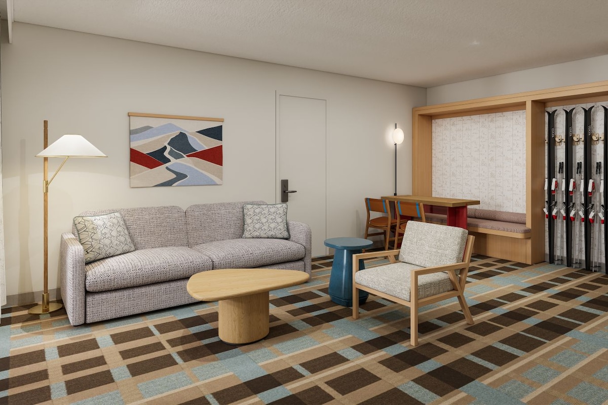 Een moderne hotelkamer met een bank, een klein rond tafeltje, een stoel, een vloerlamp, een ski-rek en een schilderij aan de muur.