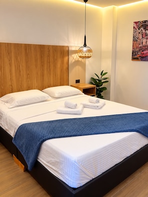 A modern hotel room with a wooden headboard, a bed with white linens, a blue throw blanket, and a hanging light fixture. - 201- A/C • Balcony • 2 Workspaces • Washing Machine • Unicentro (Medellín)