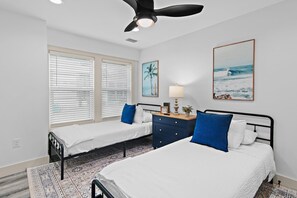 A hotel room with two beds, a ceiling fan, a window with blinds, a painting of a beach scene, and a bedside table with a lamp. - The Lookout at Betty (Santa Rosa Beach)